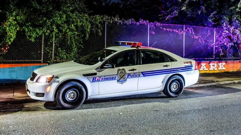 A Baltimore Police car is parked on a street at night with its lights on, next to a fence and colorful wall.