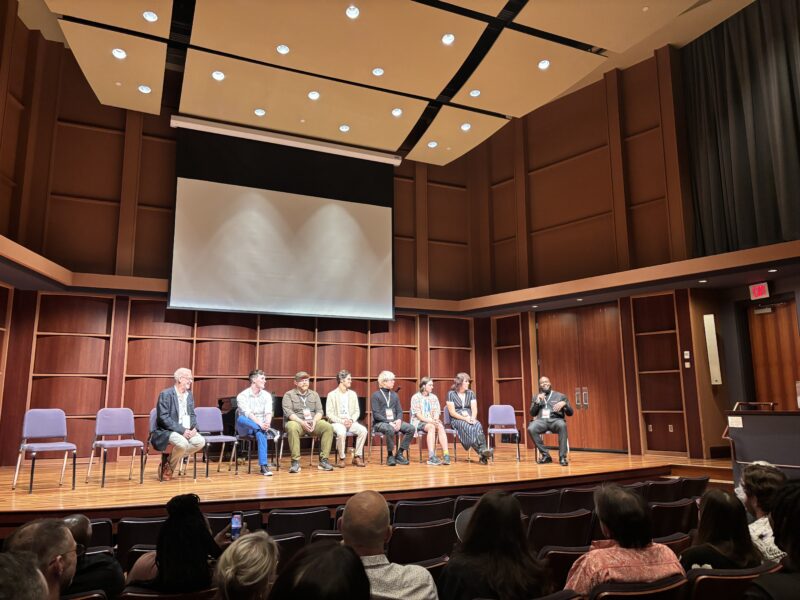 Seven people sit on stage in a wood-paneled auditorium, facing an audience. An empty podium stands to the right, and a large projection screen hangs above the panelists.