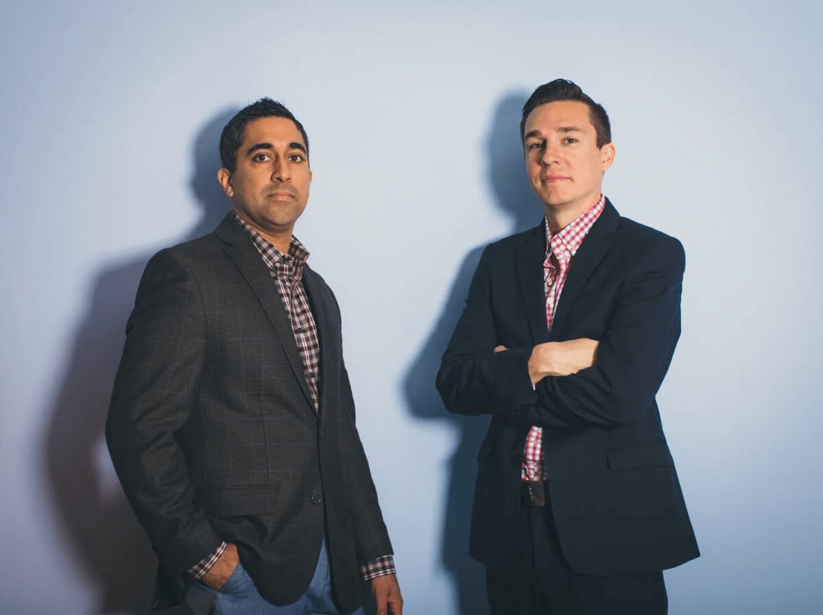 Two men in business attire stand against a plain light blue background, one with hands in pockets and the other with arms crossed, both facing the camera.