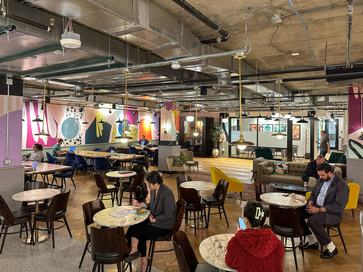 Modern coworking space with people sitting at round tables, some working on laptops or using phones, with colorful wall art and an industrial-style ceiling.
