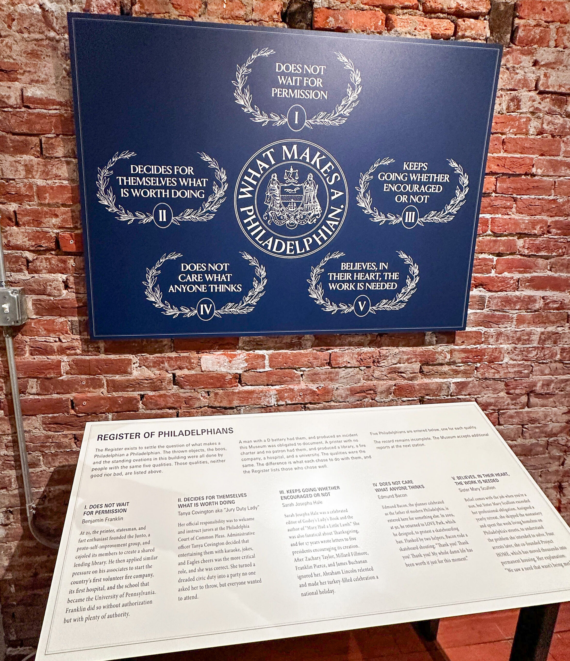 Display about “What Makes a Philadelphian” with five traits listed in a blue sign above a white plaque describing each trait, mounted on a brick wall.