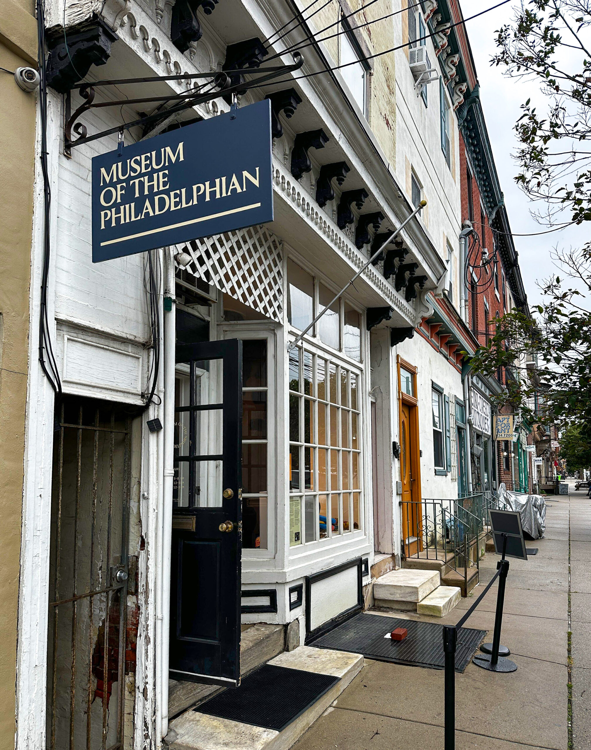 Street view of the entrance to the Museum of the Philadelphian, featuring a sign above the door and a sidewalk with a ramp and rope barrier.
