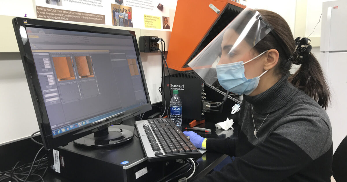 A woman wearing a face shield, mask, and gloves uses a desktop computer with laboratory equipment in the background.