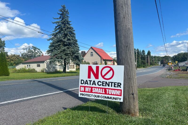 A roadside sign reads, “NO DATA CENTER IN MY SMALL TOWN! PROTECT OUR COMMUNITY,” with houses and a blue sky in the background.