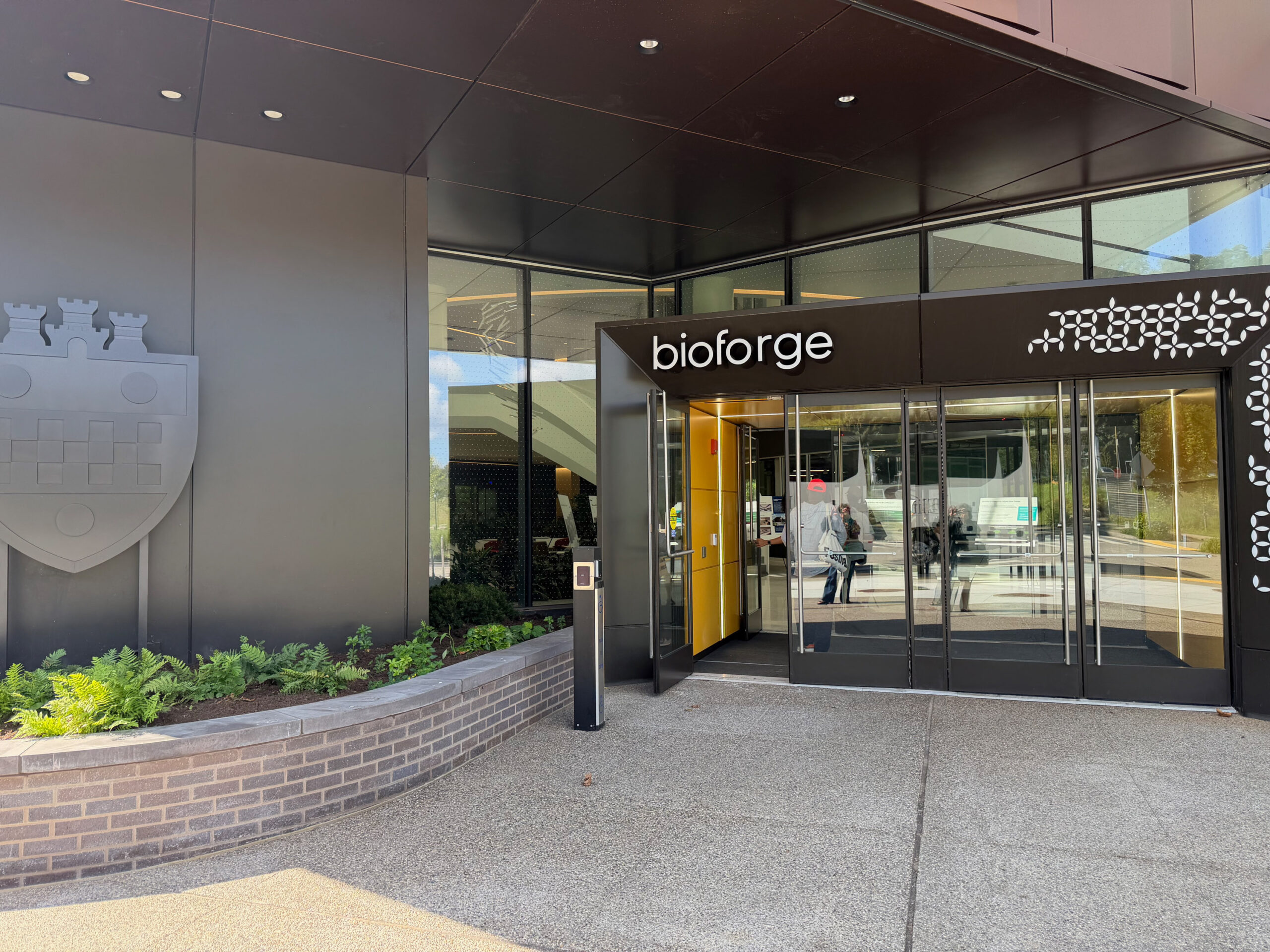 Entrance to a modern building labeled “bioforge,” with glass doors, a yellow interior wall, and a shield emblem on the exterior wall.