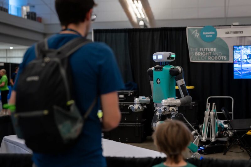 A robot stands in front of a man and a child at a robotics event booth with a sign reading, “THE FUTURE IS BRIGHT AND FULL OF ROBOTS.”.