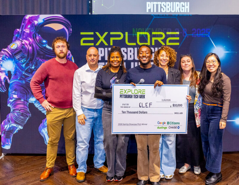 Eight people stand on stage holding a large $10,000 check for winner C.L.E.F. at the 2023 Pittsburgh Tech Week Startup Showcase. Event branding and astronaut graphic are in the background.