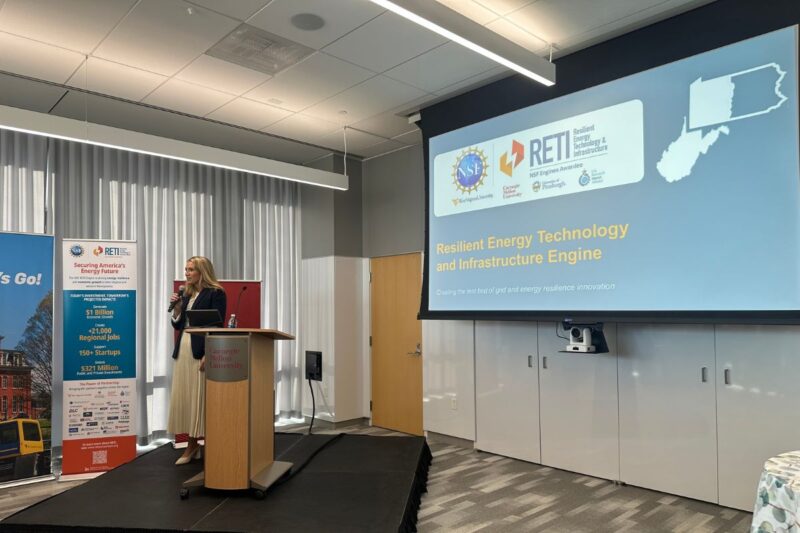 A woman stands at a podium giving a presentation on “Resilient Energy Technology and Infrastructure Engine” in a conference room with a large projection screen.