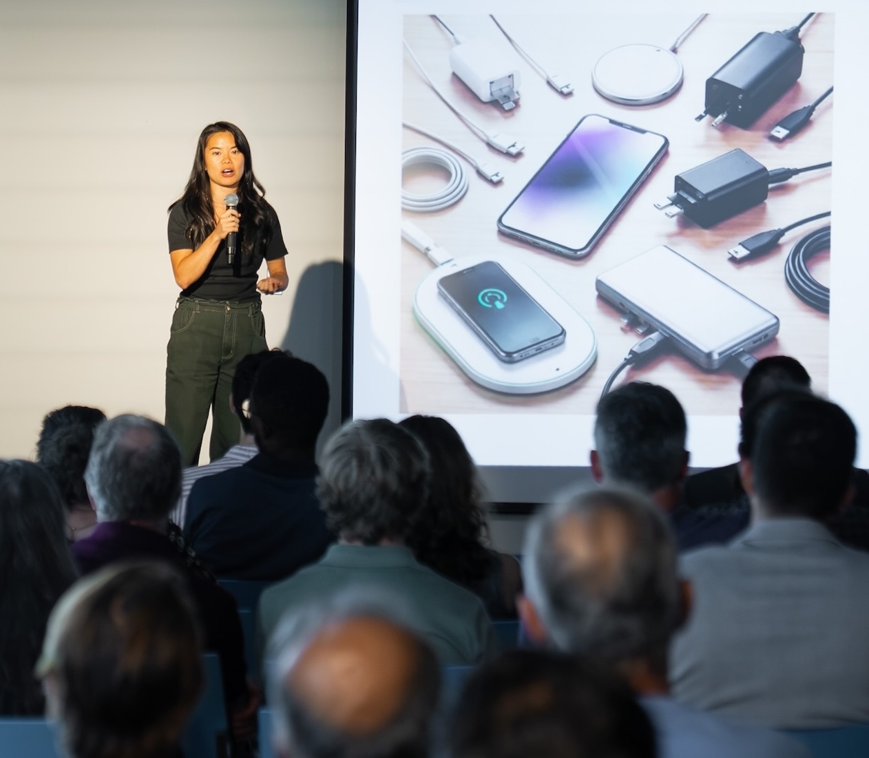 A woman speaking on stage in front of an audience, with a projected image showing various phone chargers and charging devices.
