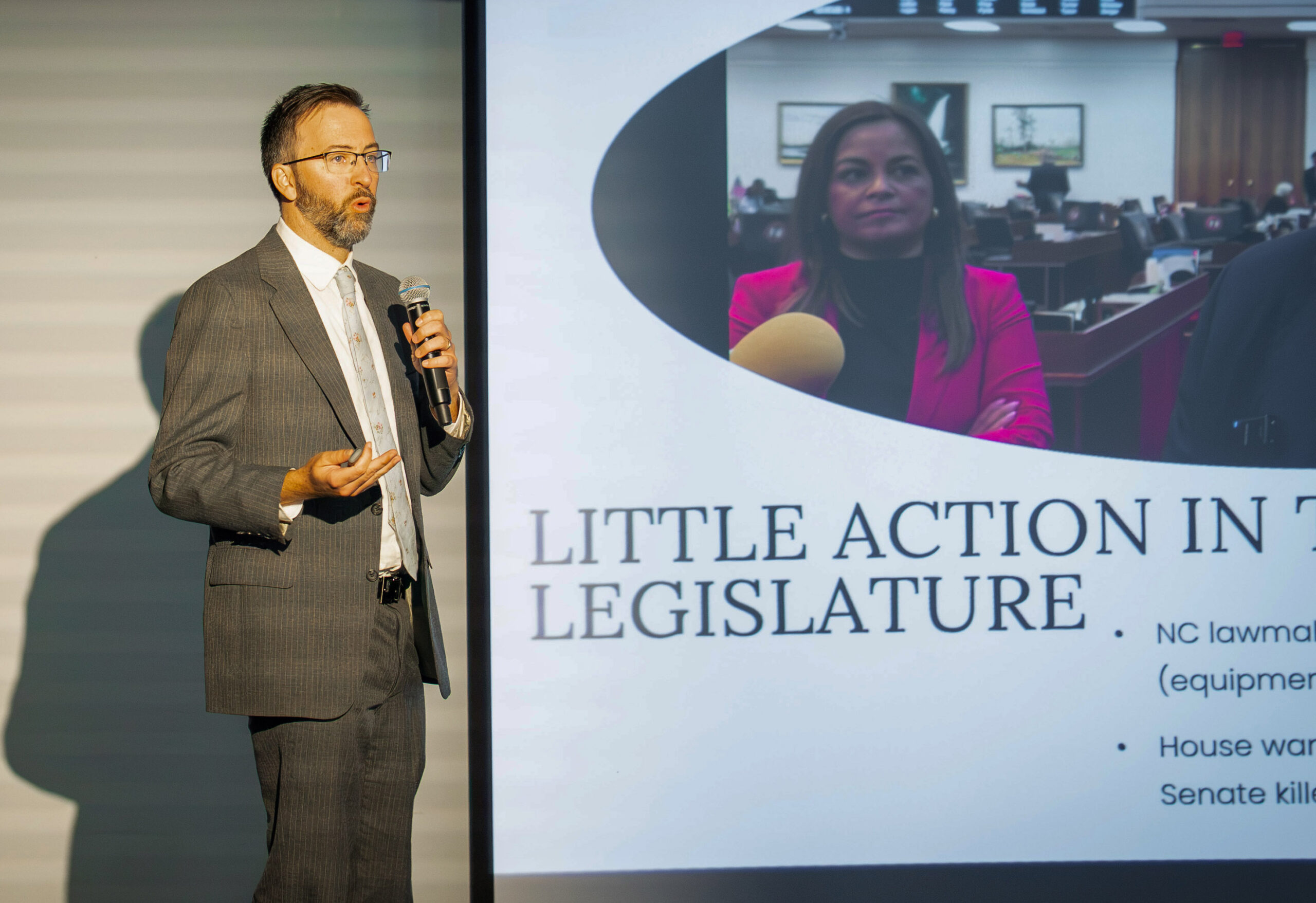 A man in a suit speaks into a microphone while standing next to a projected slide that reads, “LITTLE ACTION IN THE LEGISLATURE” with a photo of a woman and additional text.