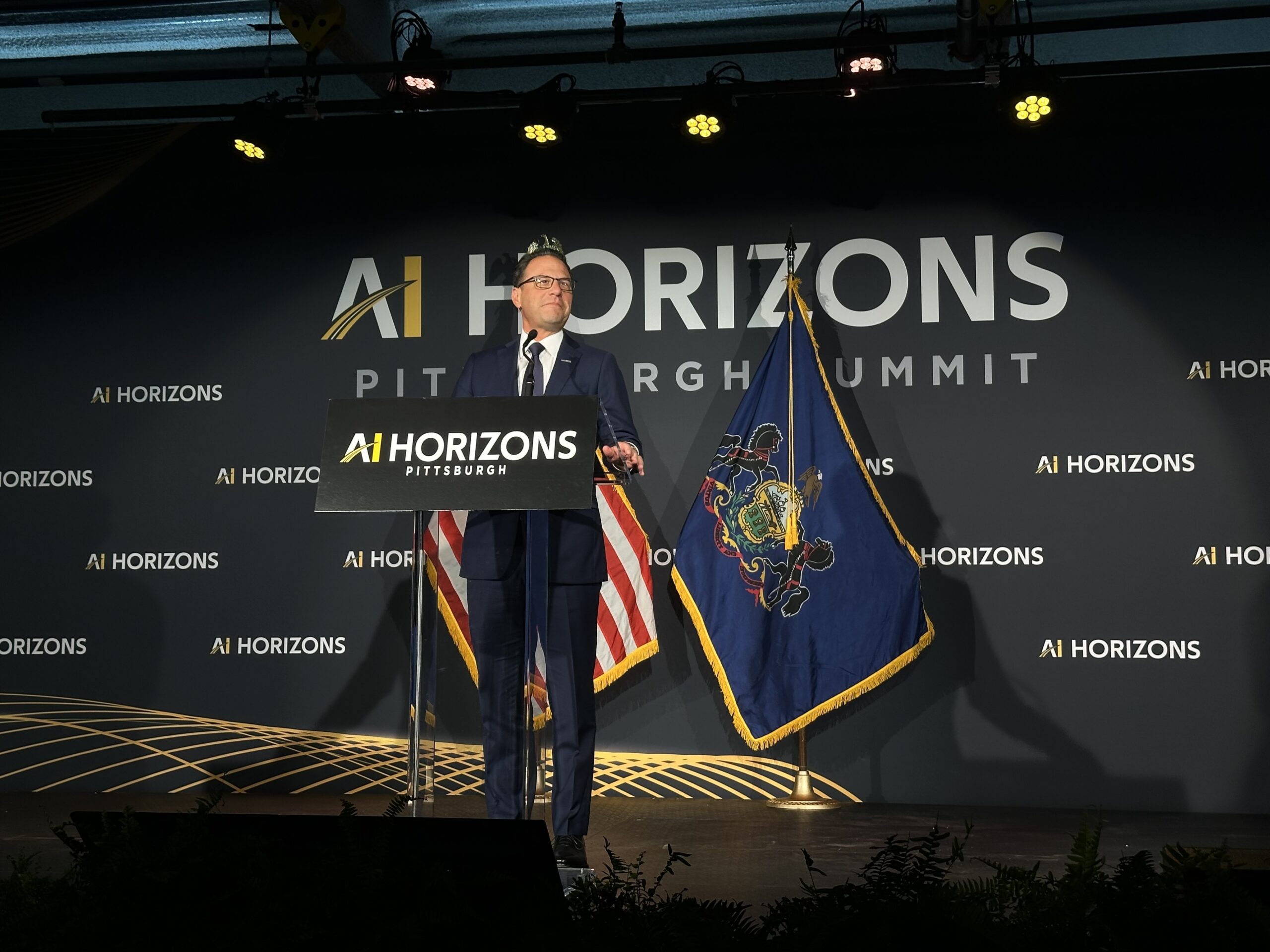 A man in a suit stands at a podium with “AI Horizons Pittsburgh” signage, flanked by U.S. and Pennsylvania flags, speaking at an event.