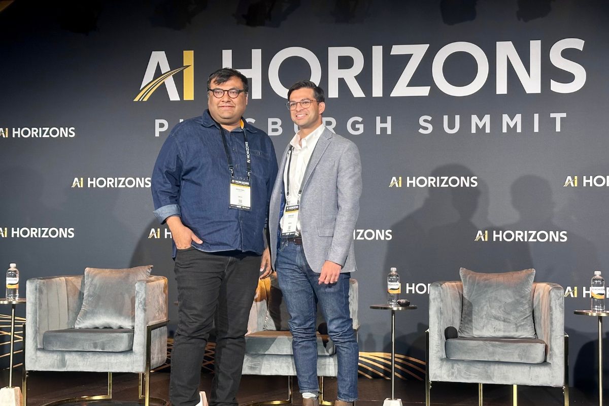 Two men stand on stage at the AI Horizons Pittsburgh Summit, posing for a photo in front of a branded backdrop and empty chairs with bottled water.