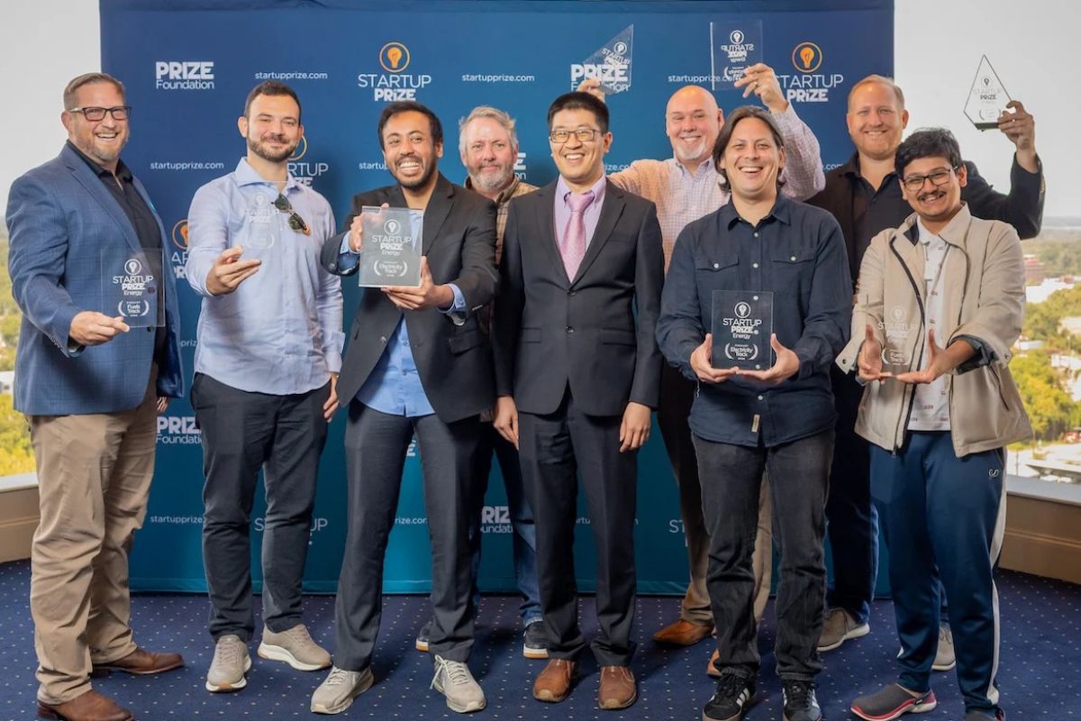 Nine people stand indoors in front of a blue “Startup Prize” backdrop, smiling and holding clear awards, dressed in business casual attire.
