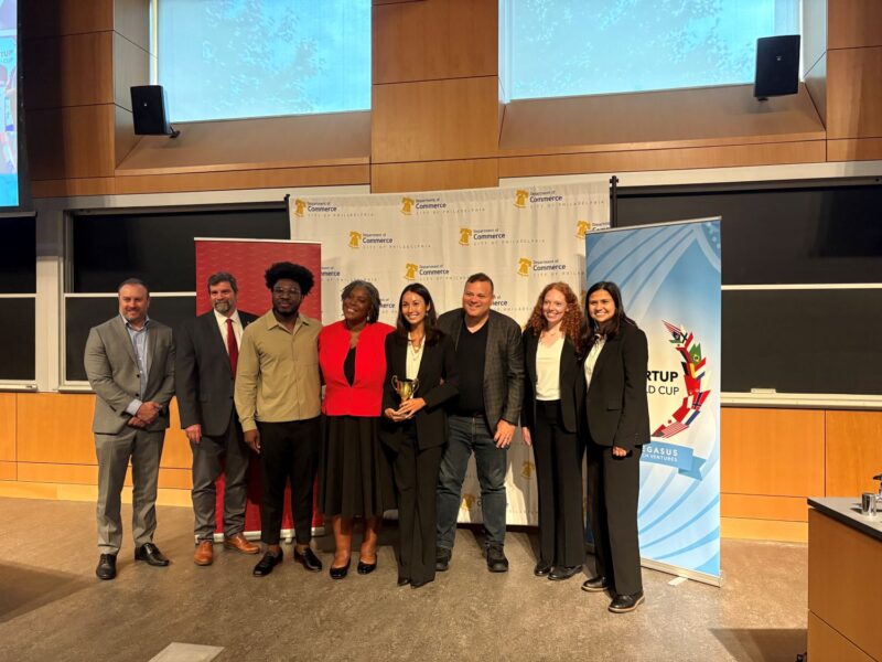 Eight people stand and pose for a group photo in front of promotional banners in a lecture hall; one woman holds a trophy.