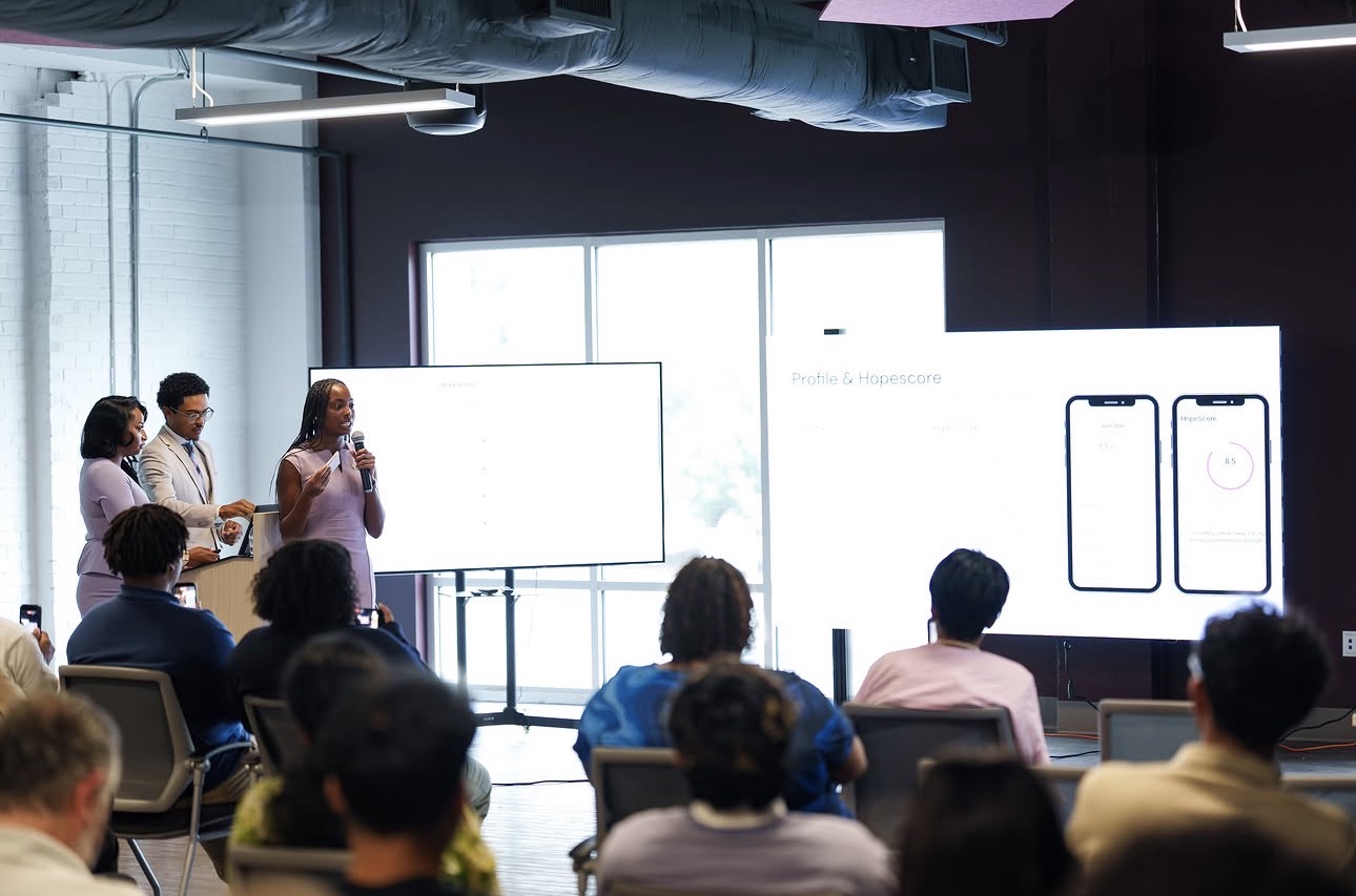 Three people present data on large screens to an audience in a modern conference room with bright windows and exposed ceilings.