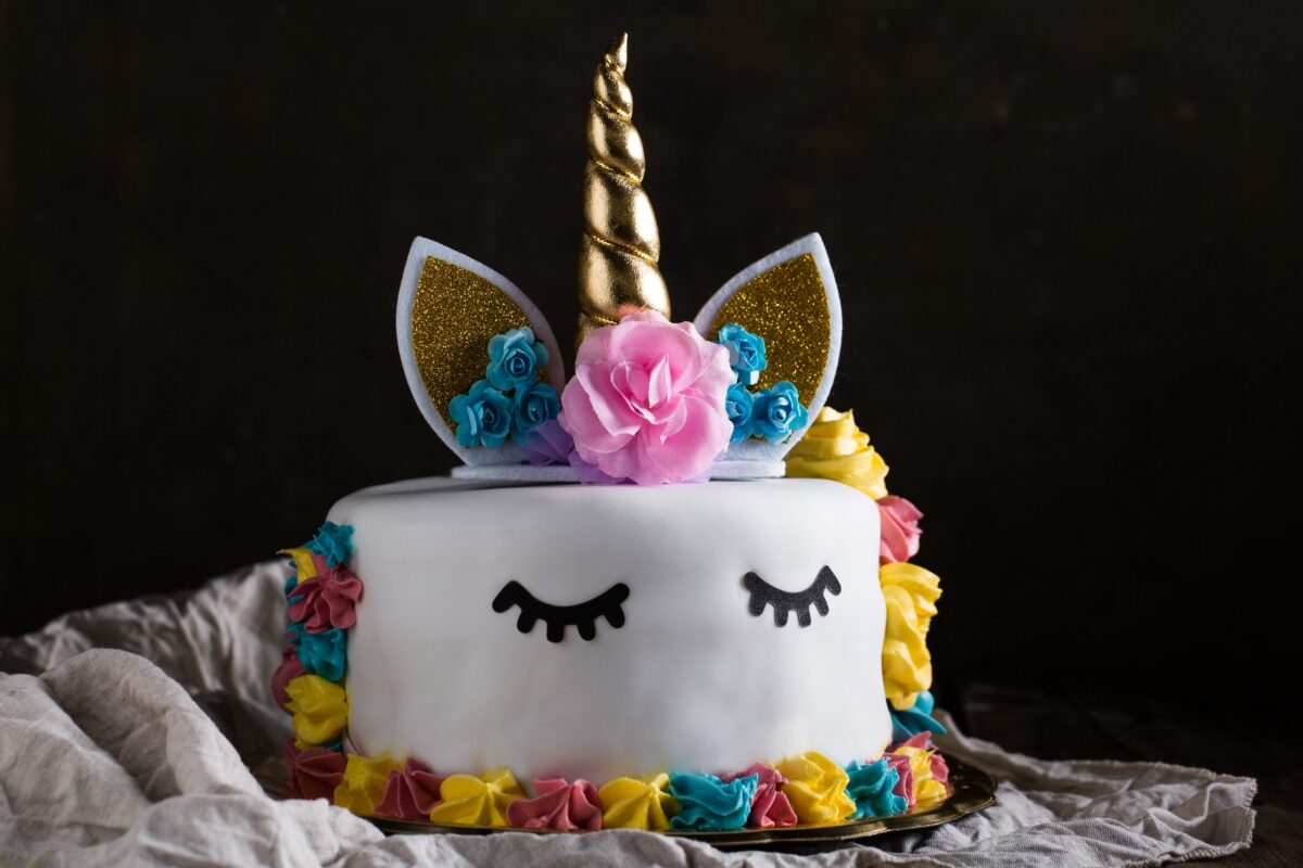 A round cake decorated as a unicorn with gold horn, ears, closed eyes, and colorful flowers on a dark background.