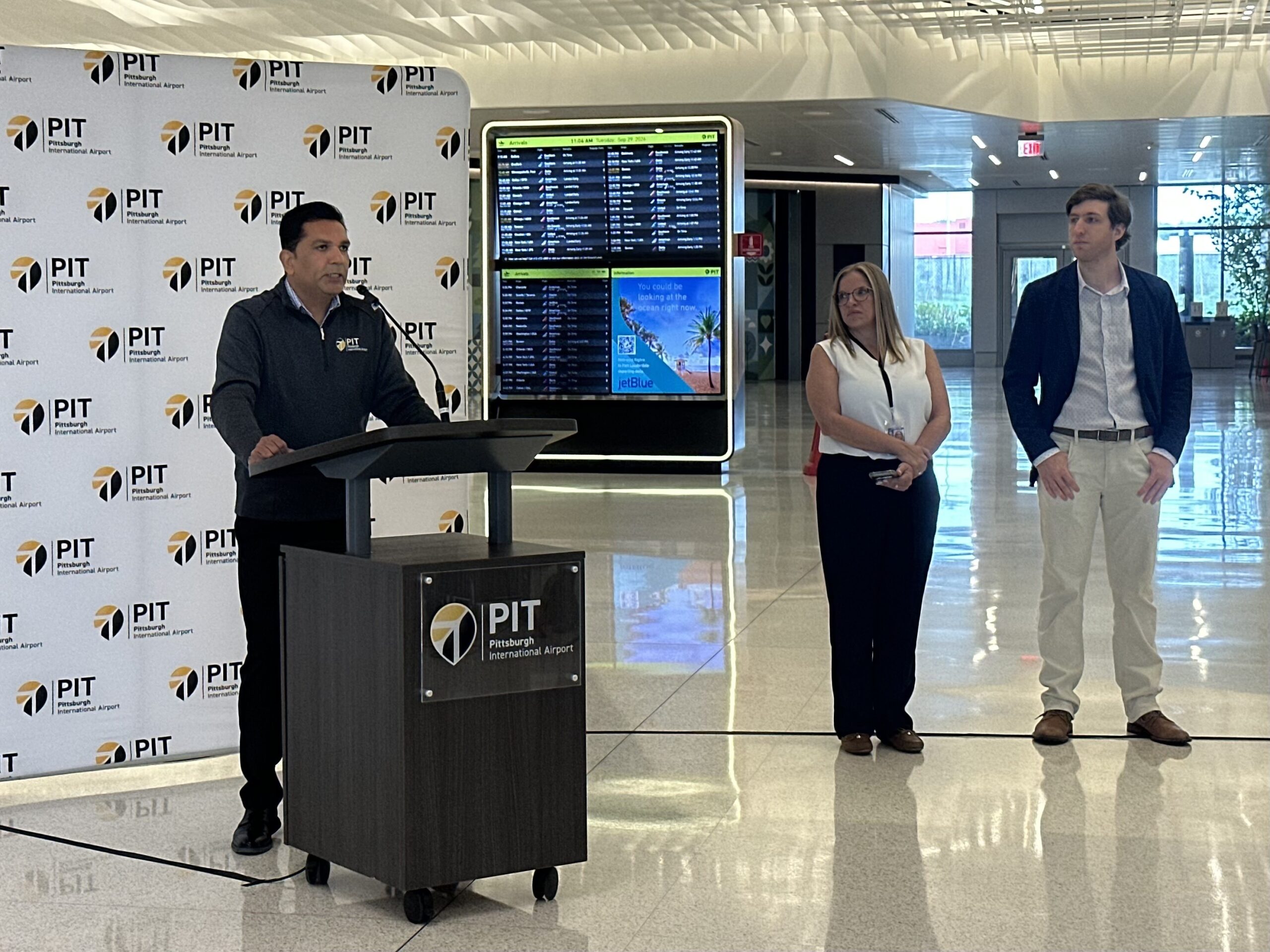 A man speaks at a podium with a PIT logo, while two people stand nearby; an airport flight information board is visible in the background.