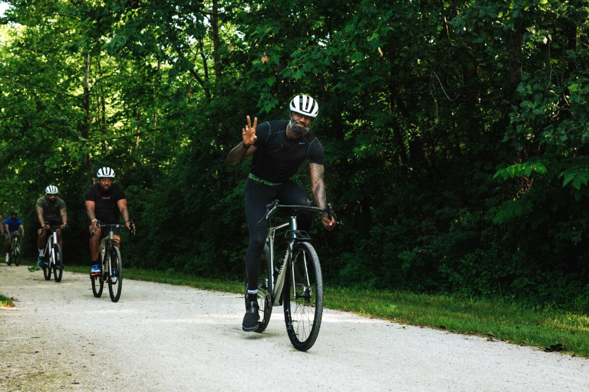 Three men wearing helmets and athletic gear ride bicycles along a wooded gravel path; the lead rider makes a peace sign with his hand.