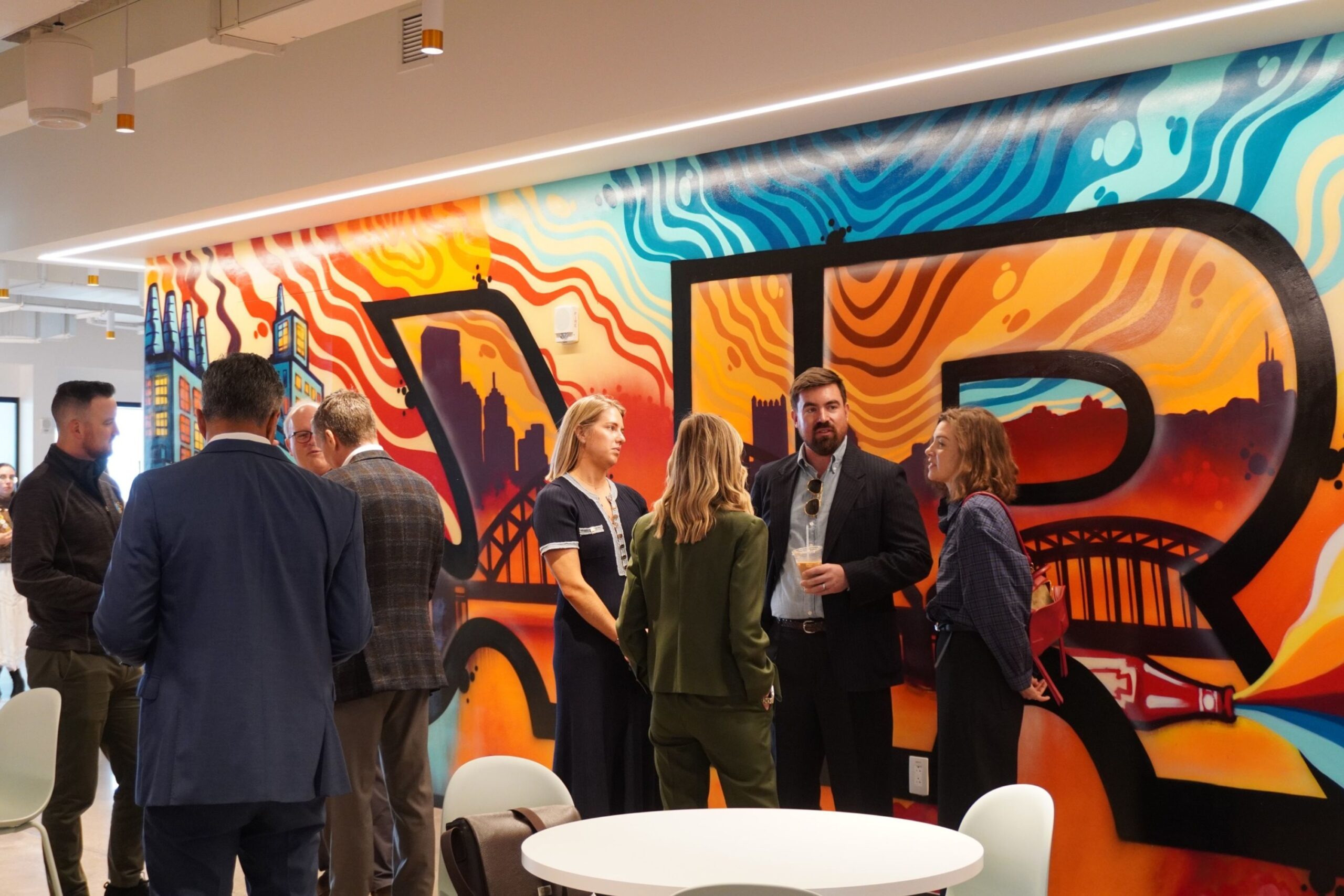 A group of people standing and talking in front of a colorful, large-scale mural of a city bridge.