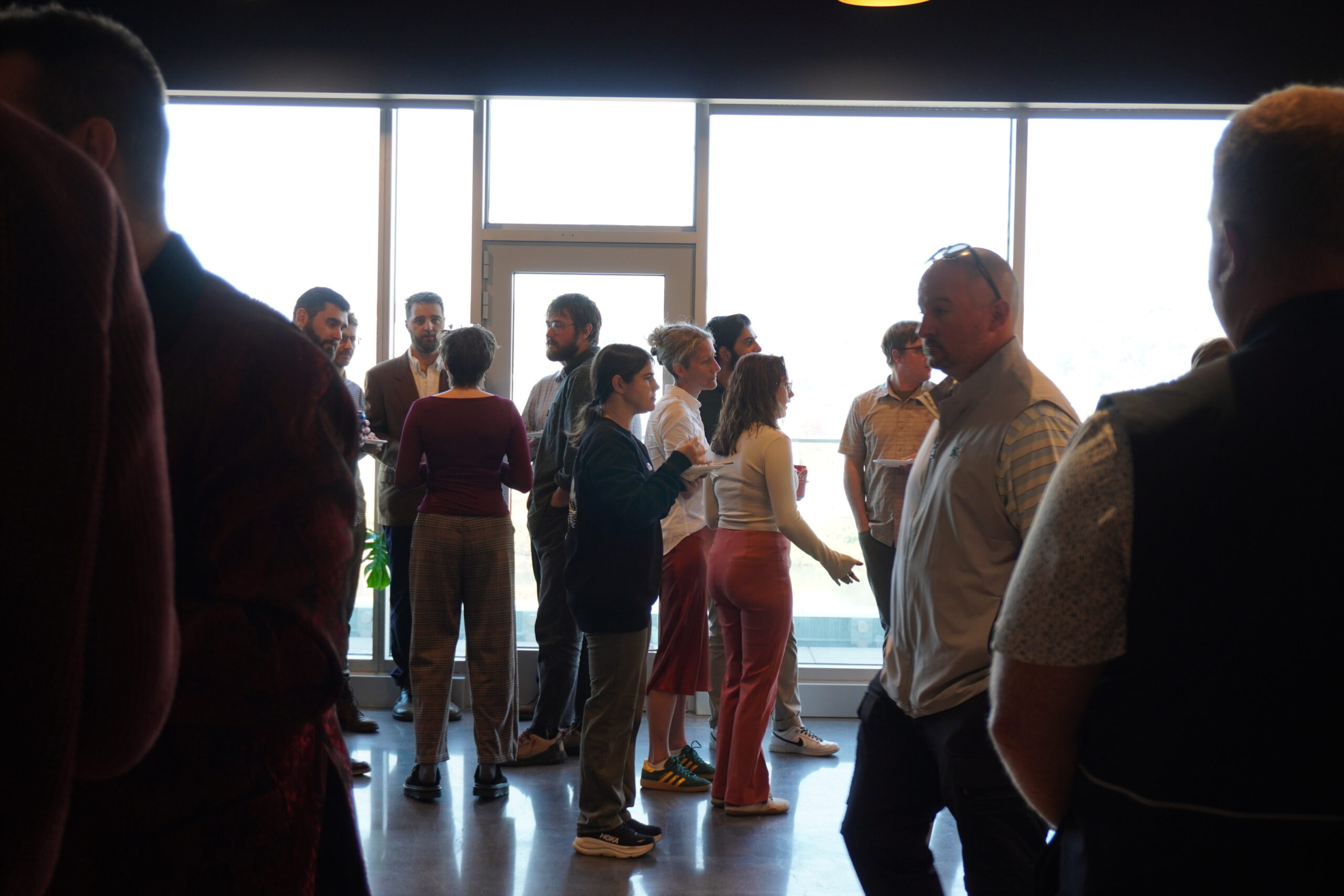 A group of people standing and talking in a room with floor-to-ceiling windows.