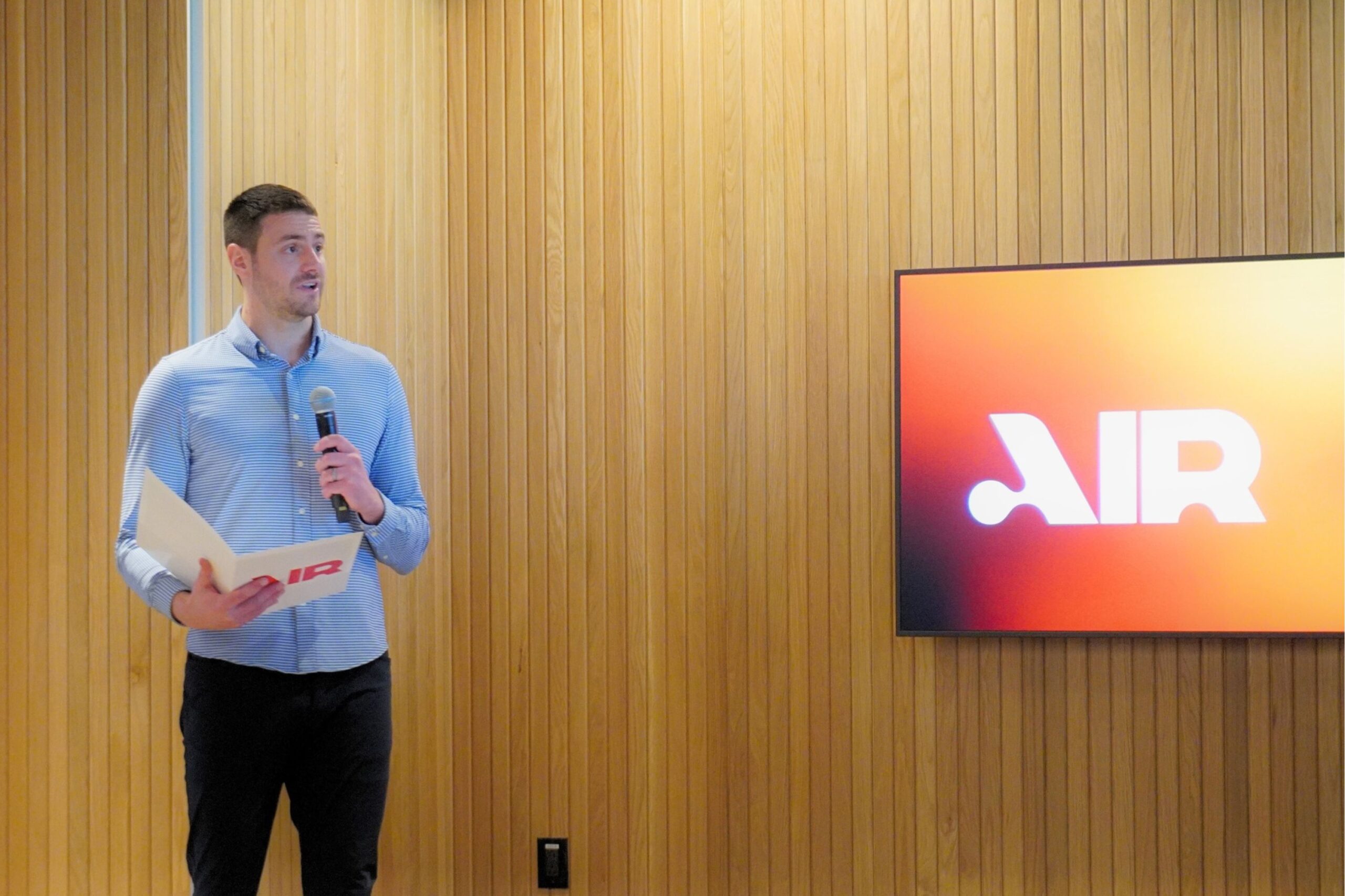 A man in a blue button-down shirt holding a microphone and a folder stands next to a screen displaying the โAIRโ logo.