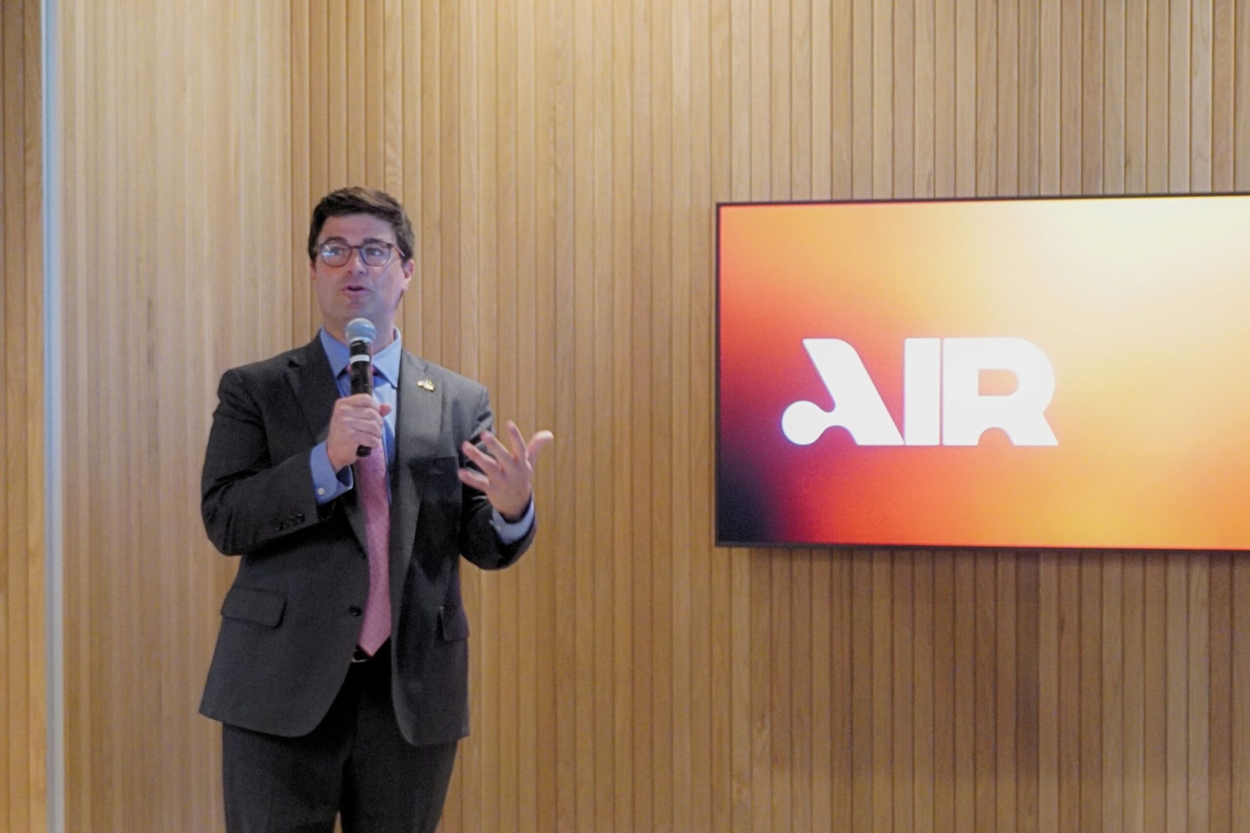 A man in a suit holding a microphone speaks in front of a screen displaying the word โAIR.โ