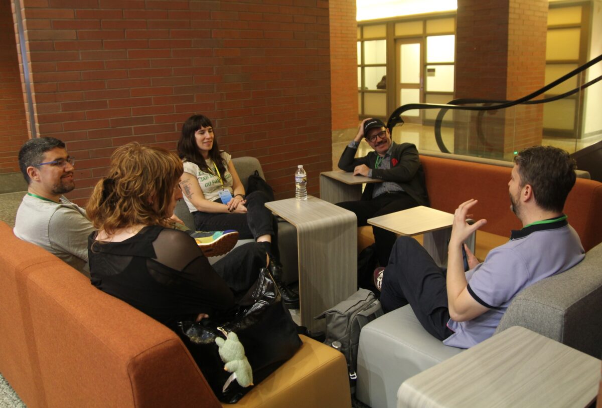 Five people sit in a lounge area with orange chairs and small tables, engaged in conversation.