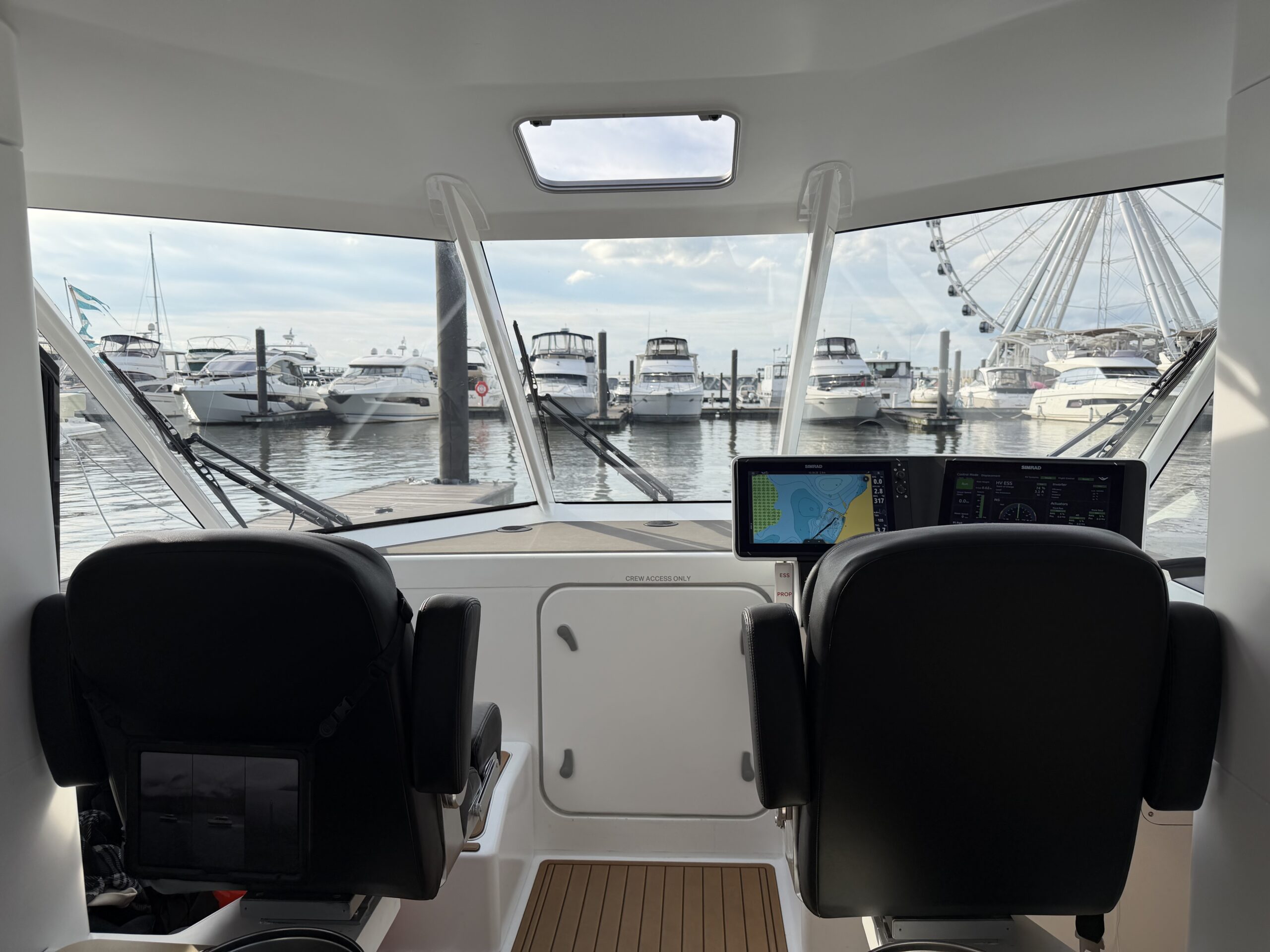 View from the helm of a boat looking out at a marina with yachts and a ferris wheel in the background.