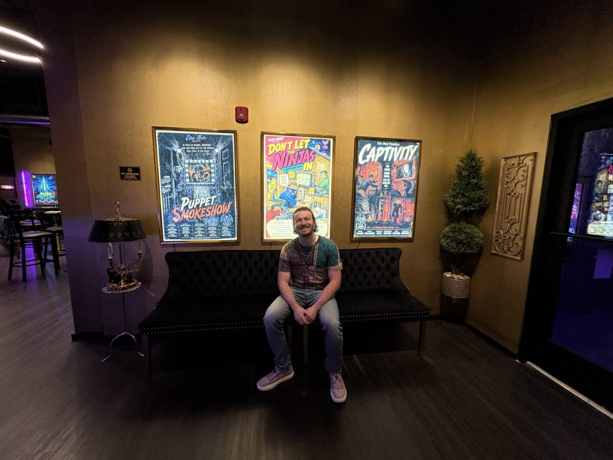 A smiling man sitting on a black bench between framed movie posters on a gold wall.