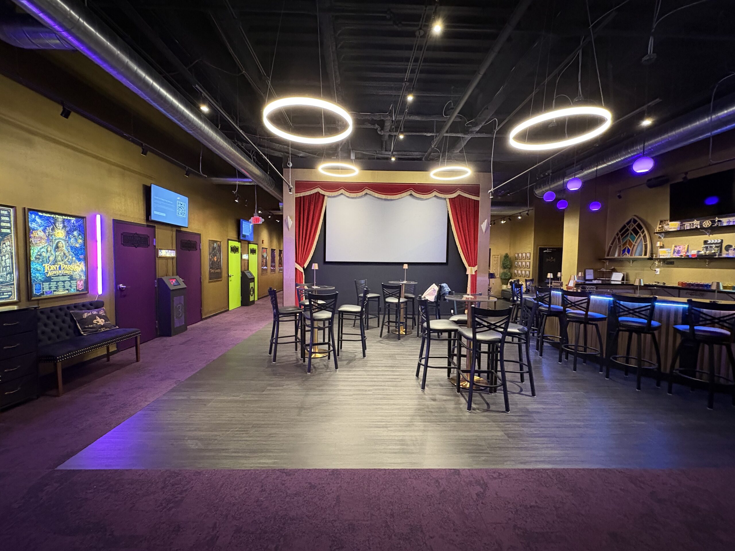 Interior of a cinema-themed lounge featuring a central stage with a movie screen, cafe-style tables, and a bar with stools.