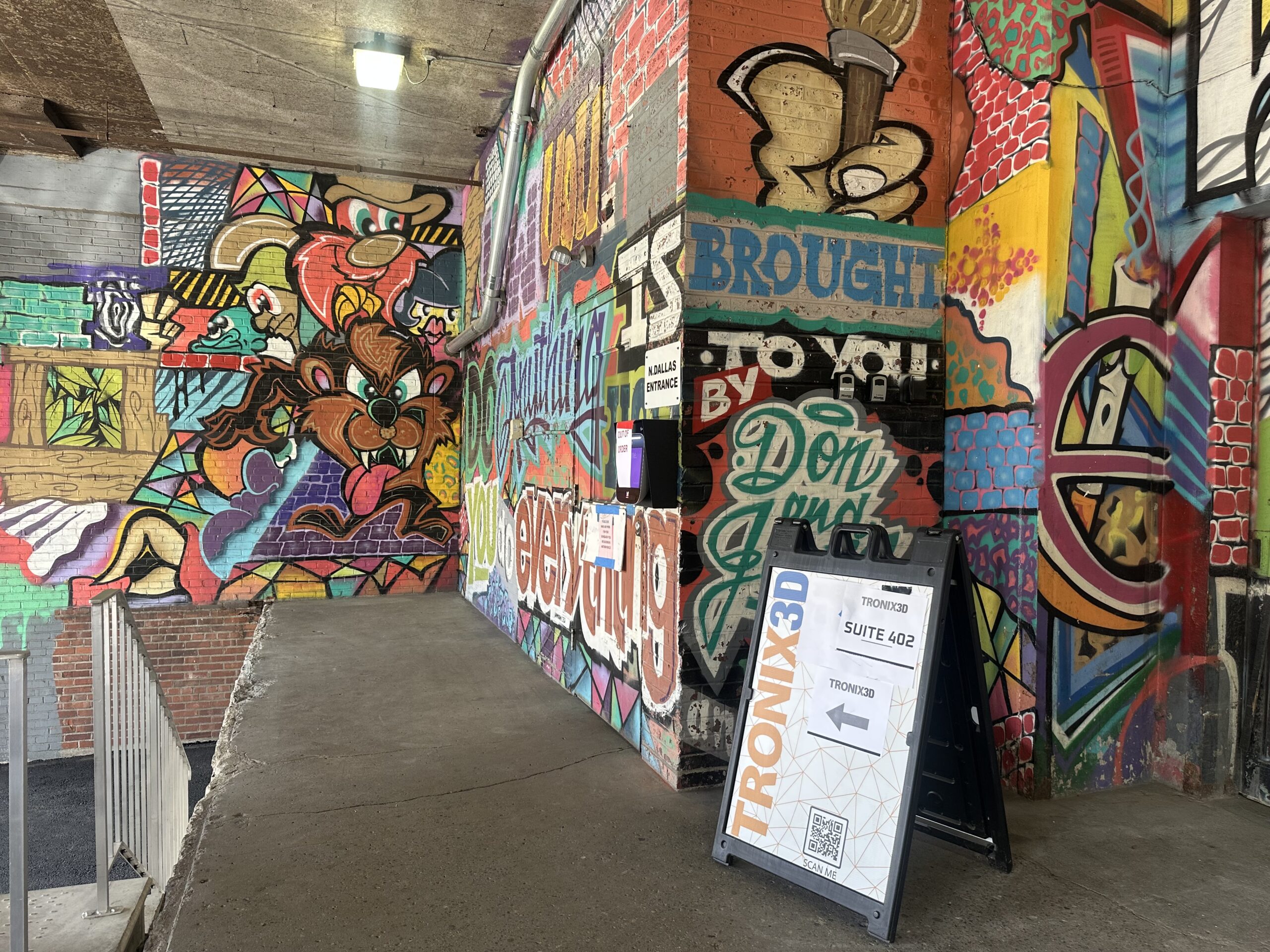 A vibrant graffiti-covered tunnel wall with a sidewalk sign directing visitors to Tronix3D, Suite 402.