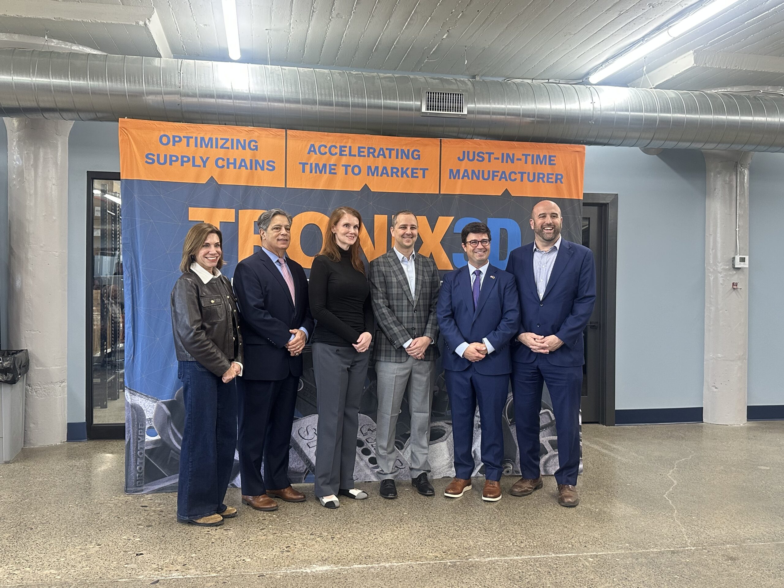 Six people stand in a line in front of a banner that reads โTRONIX3Dโ and lists the services โOptimizing supply chains,โ โAccelerating time to market,โ and โJust-in-time manufacturer.โ