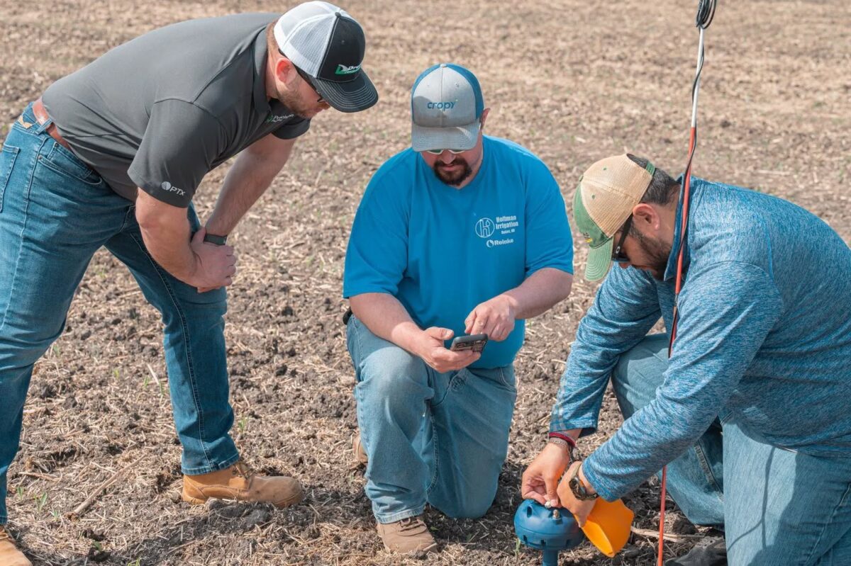 Three people crouch in a field, two watching as the third adjusts a piece of irrigation equipment and checks a smartphone.