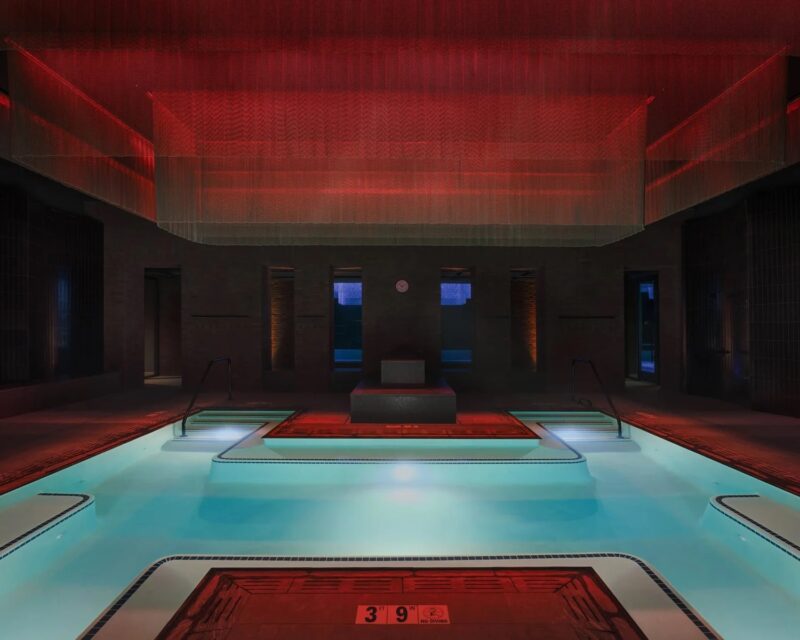 Indoor swimming pool with light blue water, illuminated by a red overhead light fixture, with a 3-foot-9-inch depth marker on the deck.
