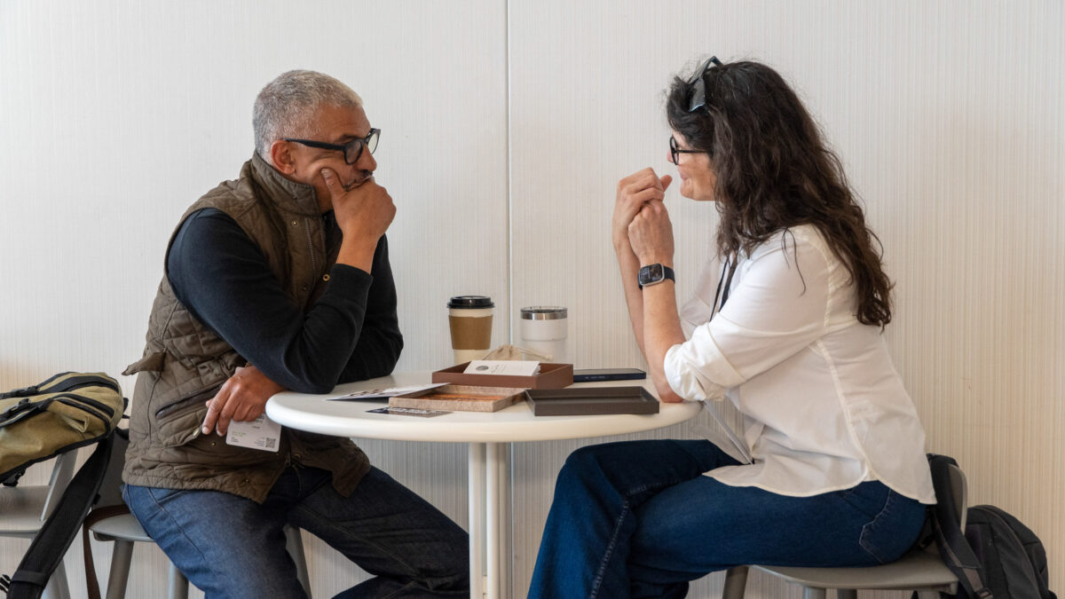 Two people in business casual attire sit at a small round table, facing each other and having a conversation.