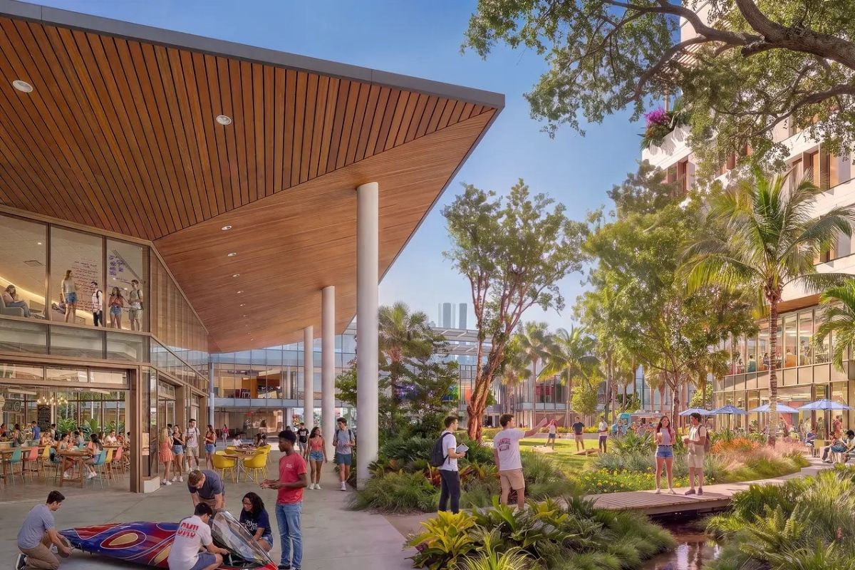 A sunny outdoor campus plaza with students walking, sitting, and socializing under a large wooden architectural overhang and near a landscaped garden with a path.