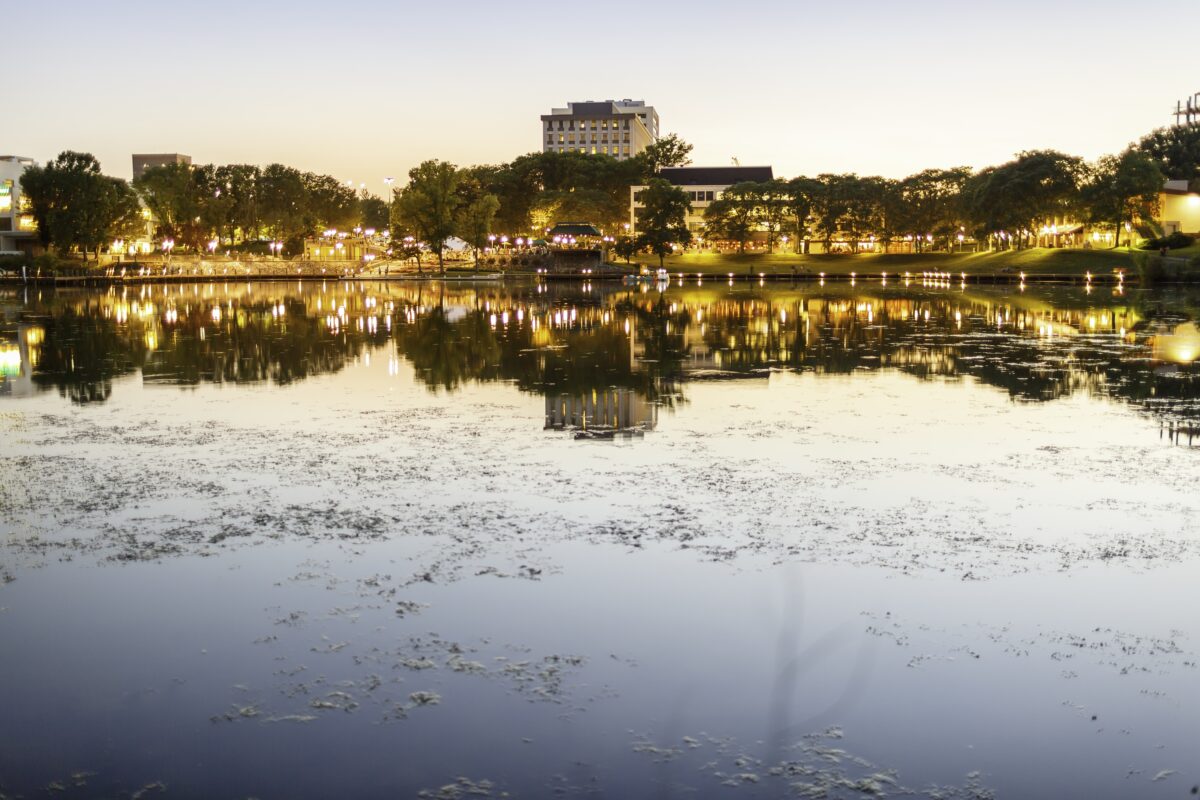 A city park and buildings at dusk reflected in a calm pond.
