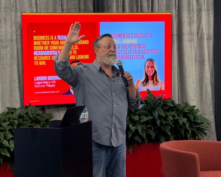 A man with a gray beard speaking into a microphone with his hand raised, standing in front of a presentation screen about Landon Donovan.