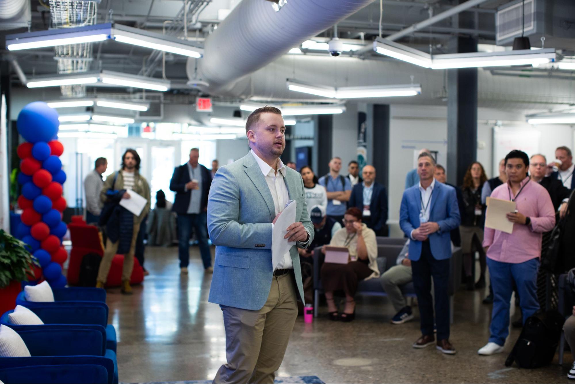 A man in a light blue blazer stands in the center of an office space, holding papers while addressing a group of people.