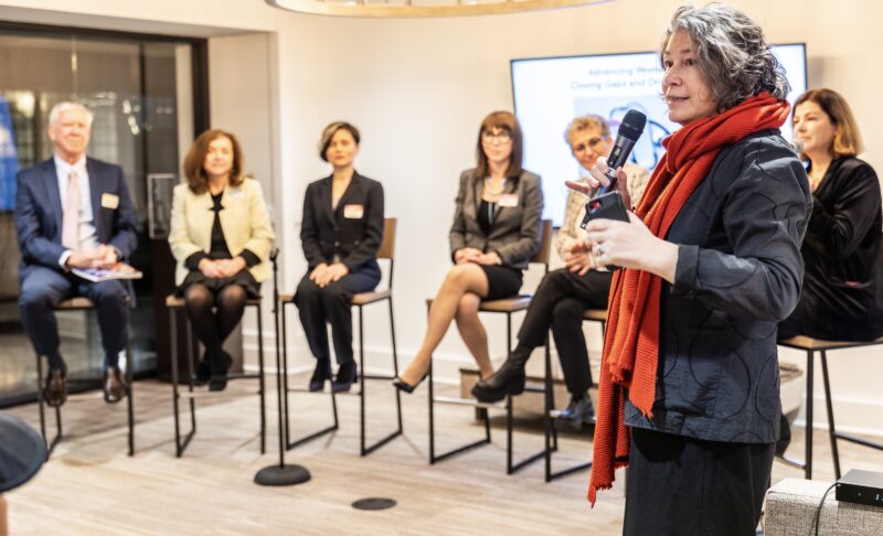 A woman speaks into a microphone at a panel discussion with five other people seated behind her.