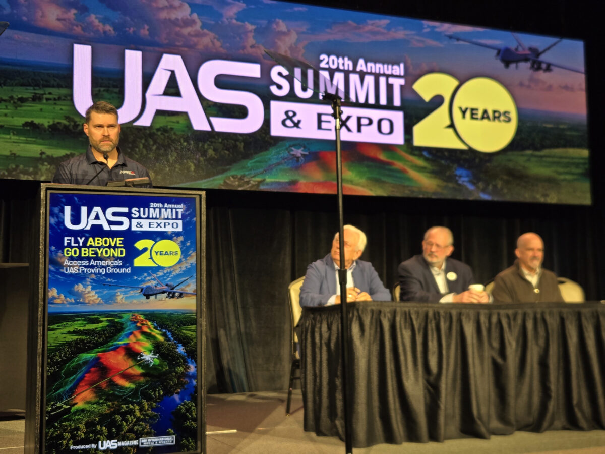 A speaker stands at a podium at the 20th Annual UAS Summit & Expo, with three panelists seated at a table on stage behind him.