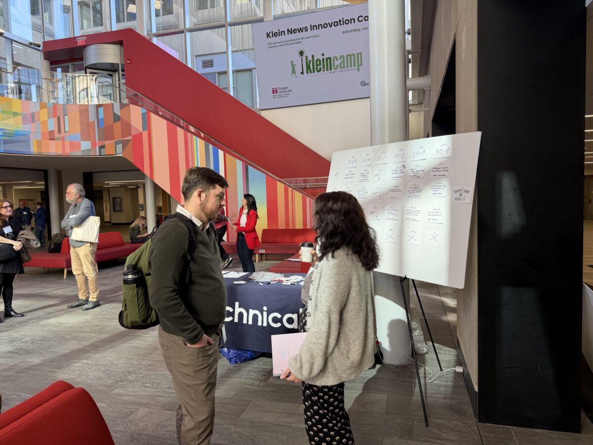Two people converse in an atrium in front of a whiteboard covered in sticky notes, with a large banner for Klein News Innovation Camp hanging above.