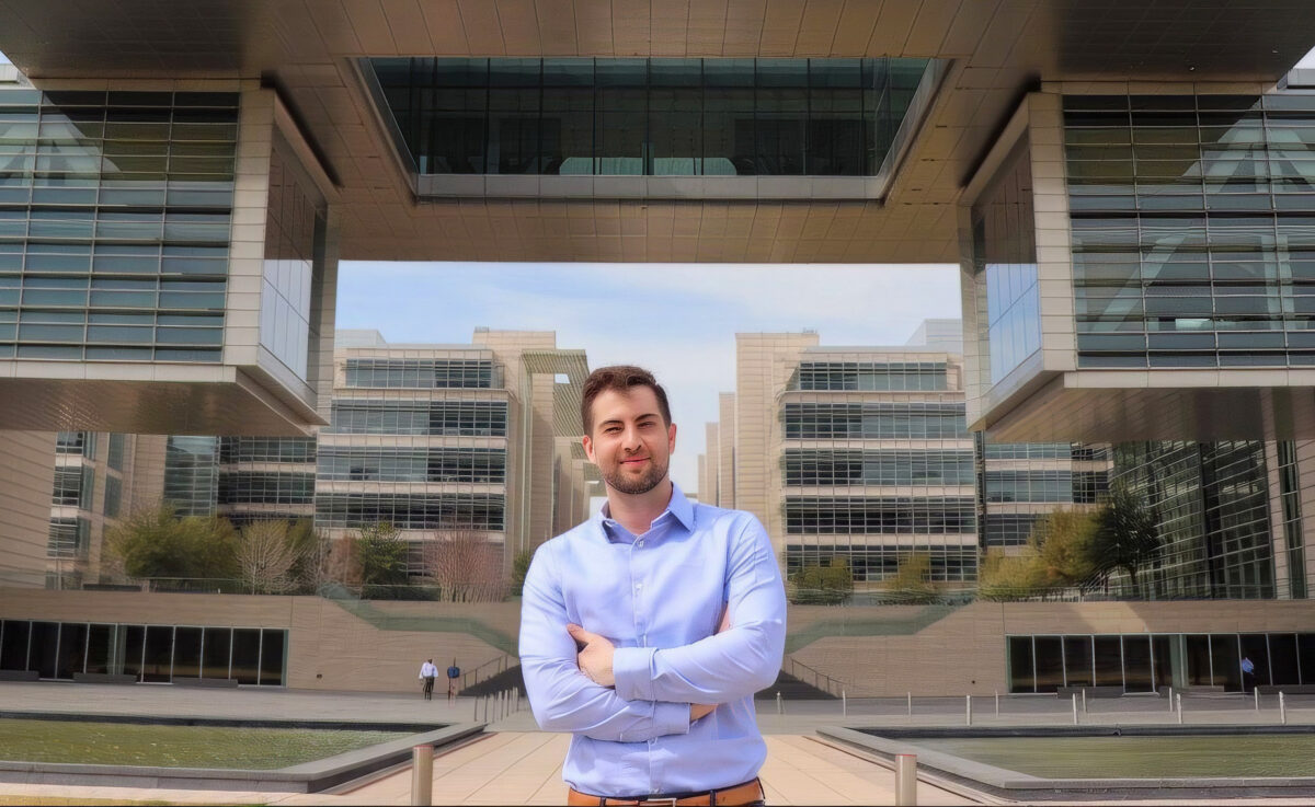 A man in a light blue button-down shirt standing with arms crossed in front of a modern office complex with a large central courtyard.