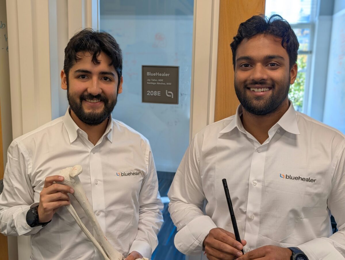 Two men in branded white shirts standing in front of an office door, one holding a model of a human tibia.