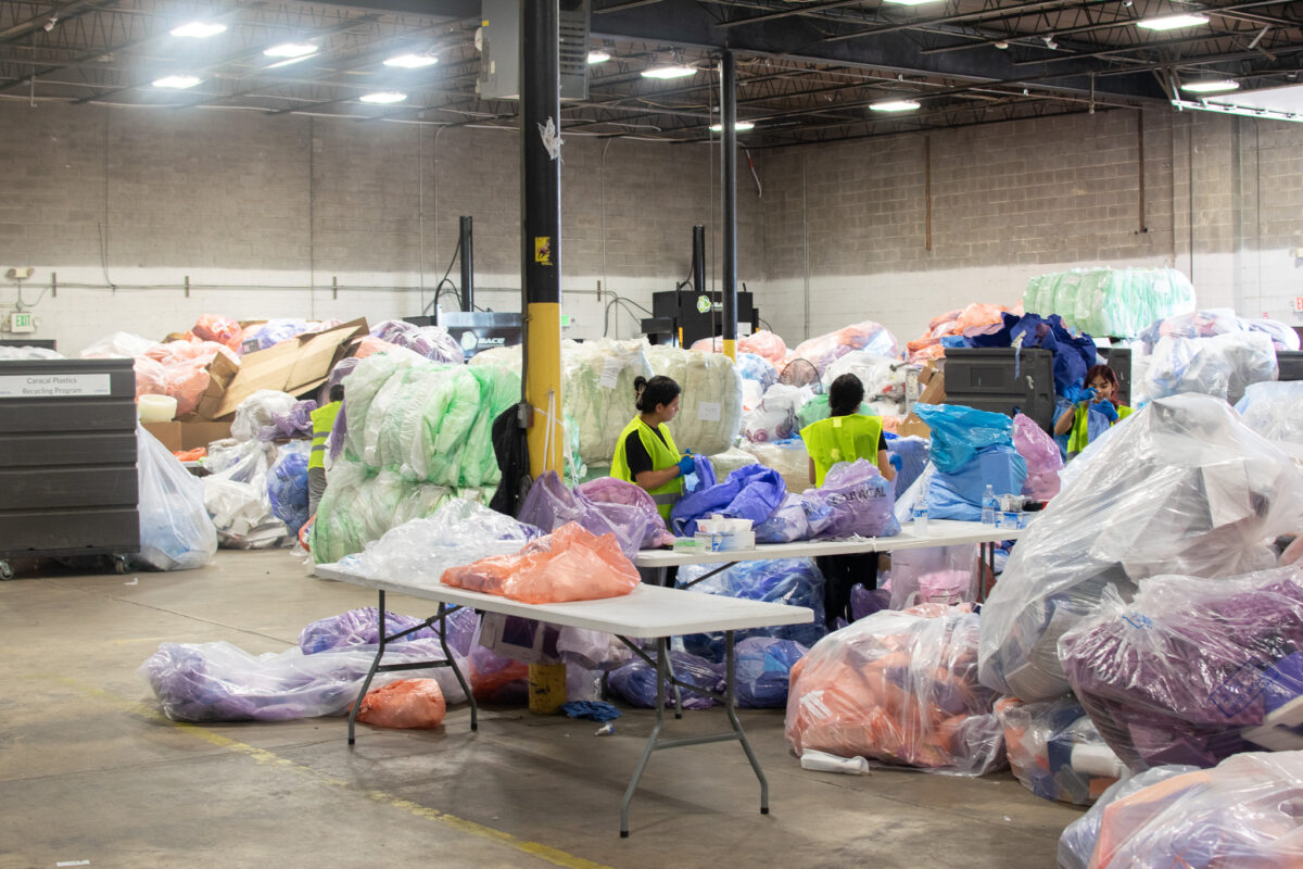 Workers in high-visibility vests sort bags of plastic waste at a recycling facility.