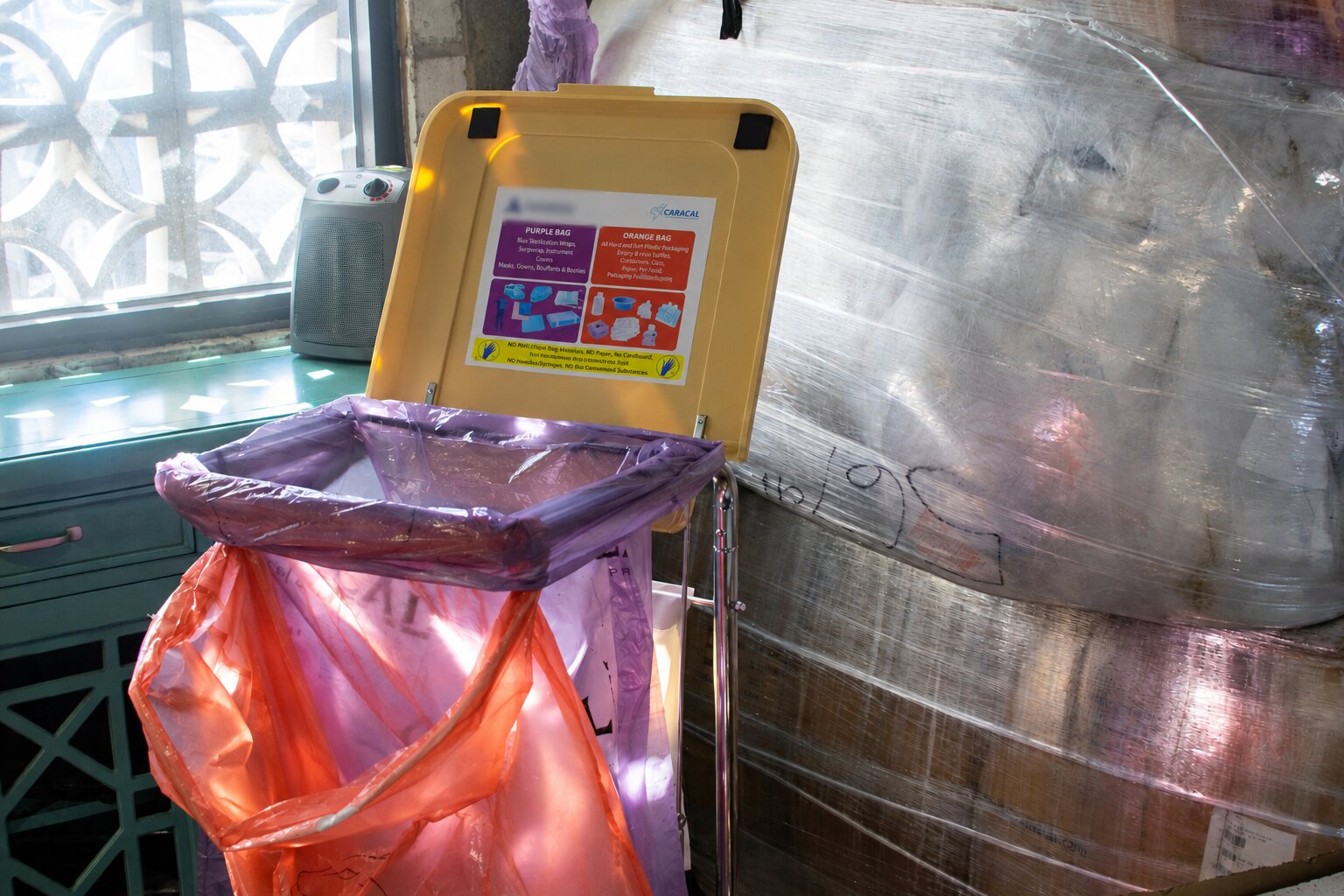 A waste sorting station with a yellow lid and two hanging bags, one purple and one orange, featuring a sign detailing proper disposal for each color.