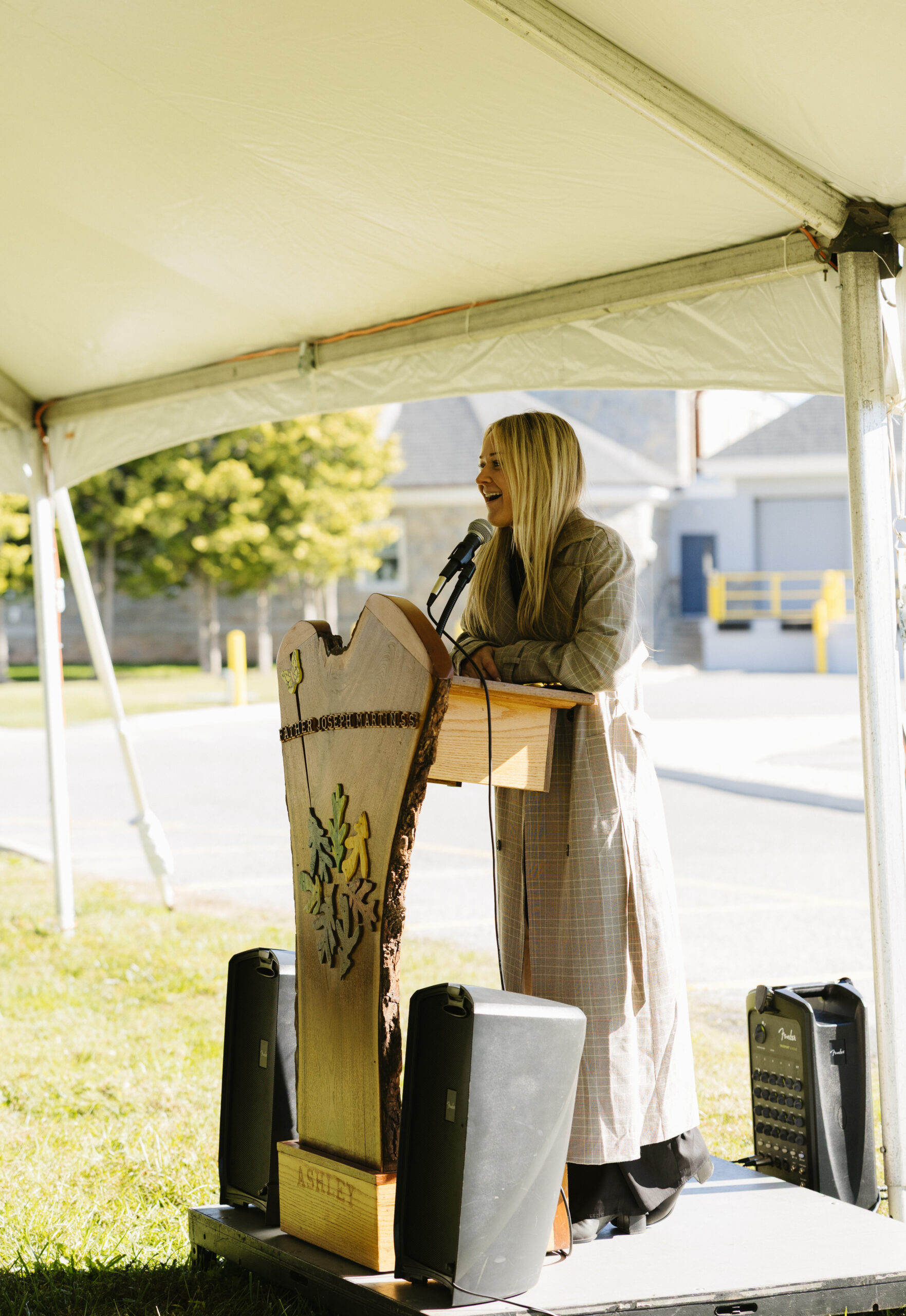 A woman stands at a wooden podium speaking into a microphone under a white tent.