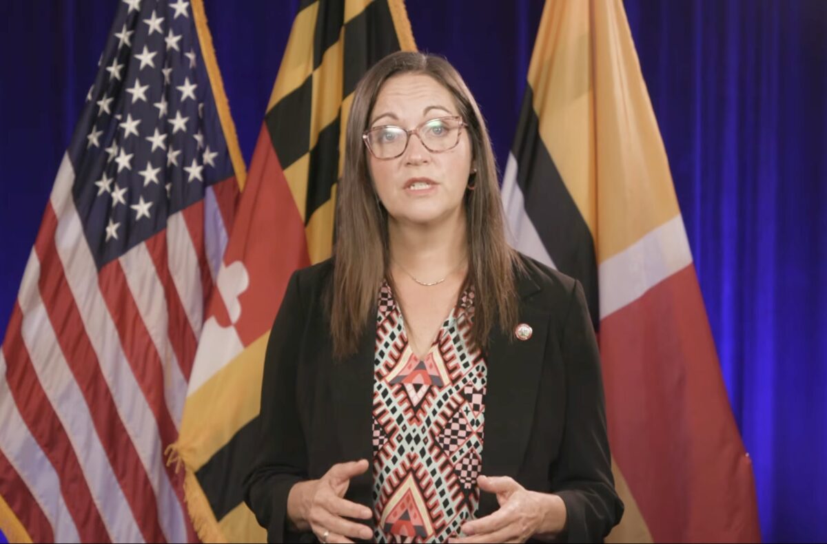 A woman with glasses and a black blazer speaks in front of the United States and Maryland flags.