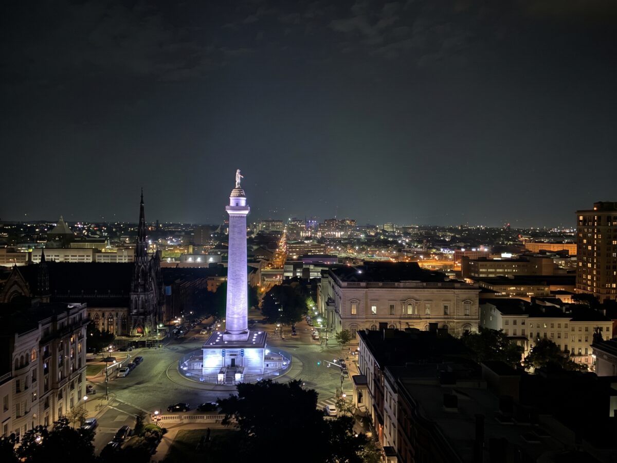 The Washington Monument in Baltimore illuminated at night, surrounded by historic buildings and city streets.
