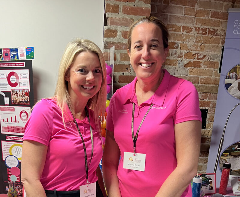 Two women in matching pink polo shirts and name tags pose for a photo at a display booth.